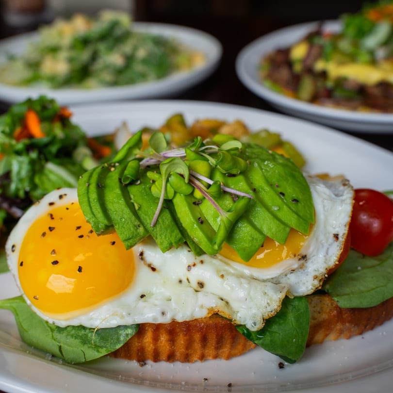 Avocado toast with two fried eggs, spinach, and cherry tomatoes, served with a side salad.