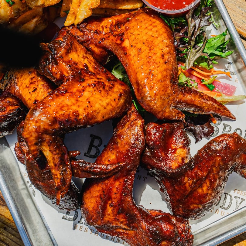Close-up of Brody's BBQ chicken wings with fries, salad, and dipping sauce on a metal tray.