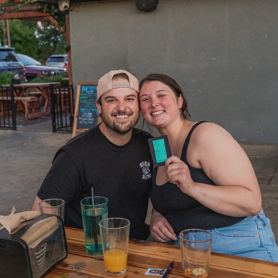 A smiling couple, a man in a cap and a woman holding a green card, sitting at a Brody's BBQ outdoor table.