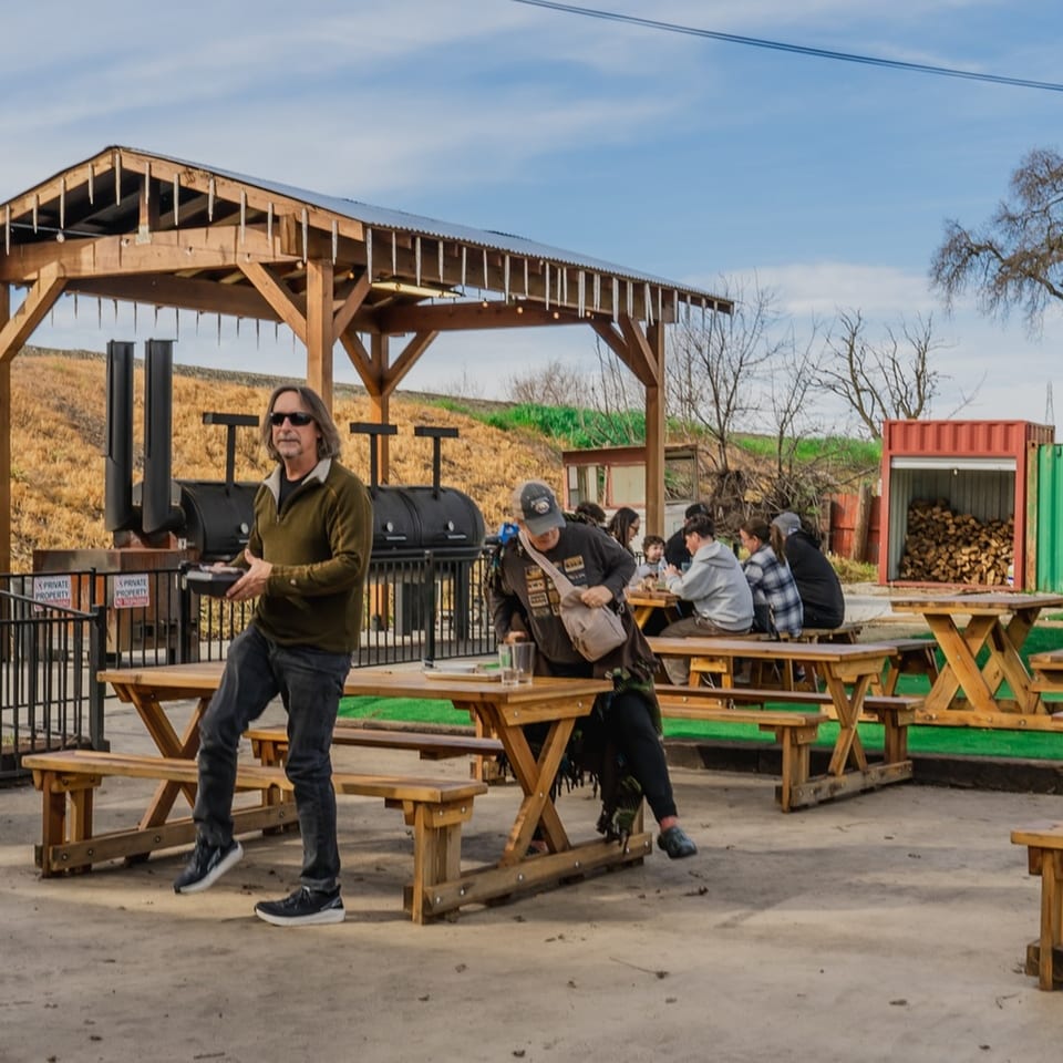 Outdoor BBQ restaurant patio with picnic tables, people dining, and large smokers under a wooden canopy.
