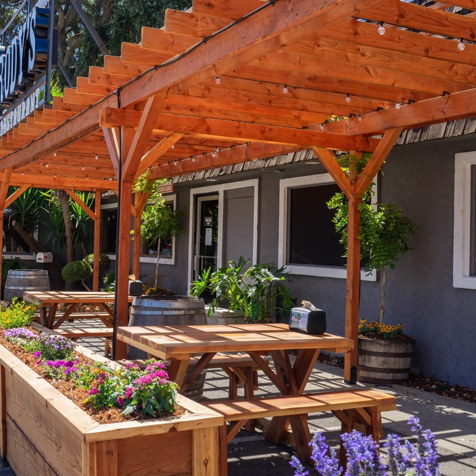 Brody's Restaurant outdoor patio with wooden picnic tables under a pergola, surrounded by plants and flowers.