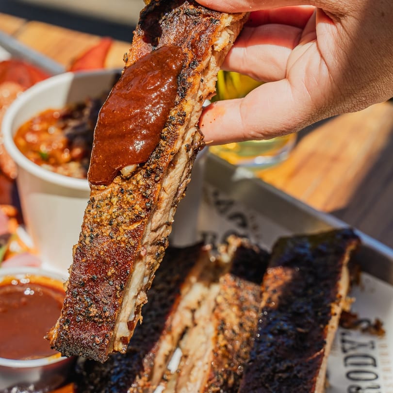 A hand holds up a saucy, seasoned Brody's BBQ rib, with more ribs and sides blurred in the background.
