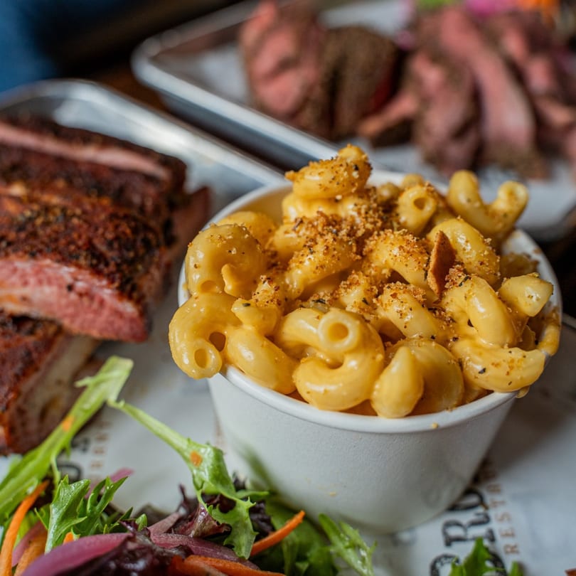 Brody's BBQ mac and cheese in a white cup, with ribs and salad in the background.