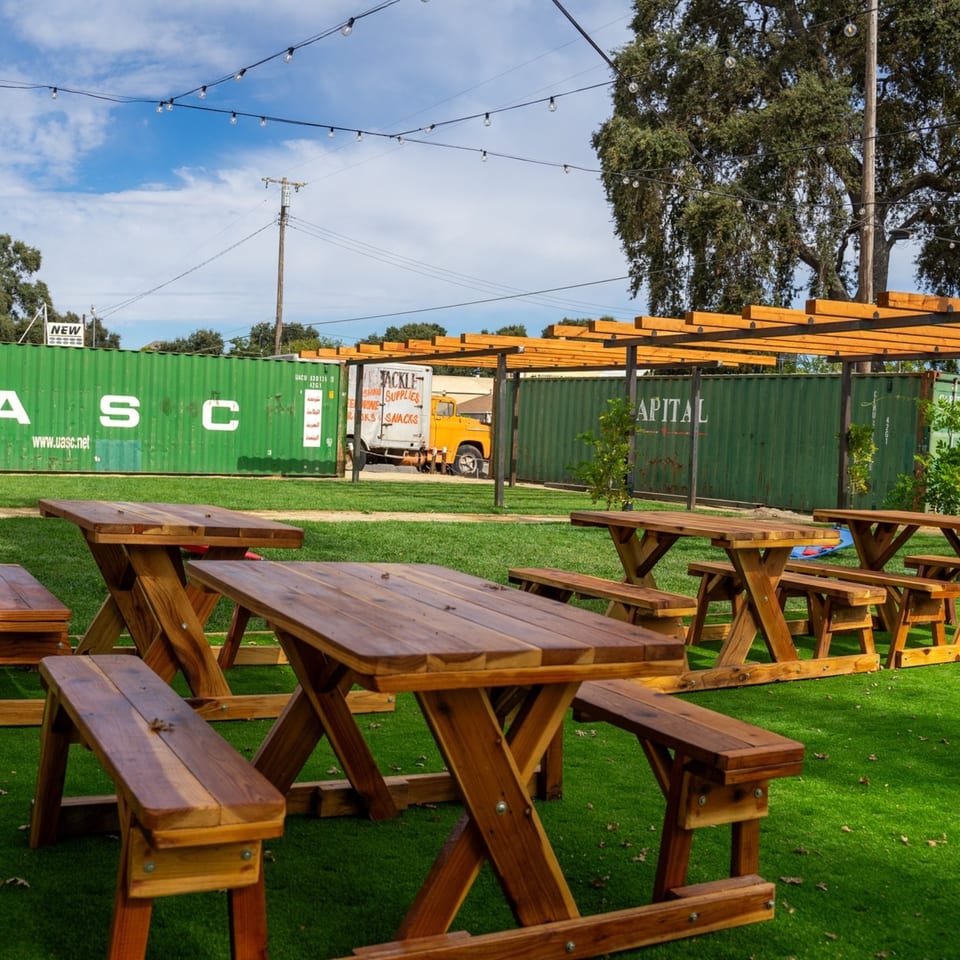 Outdoor seating area with wooden picnic tables on artificial grass, string lights overhead, and green shipping containers.